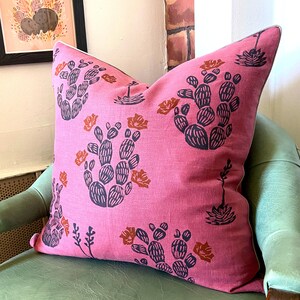May include: A pink decorative pillow with a cactus pattern in navy blue and orange. The pillow is on a green chair, with a framed picture in the background.
