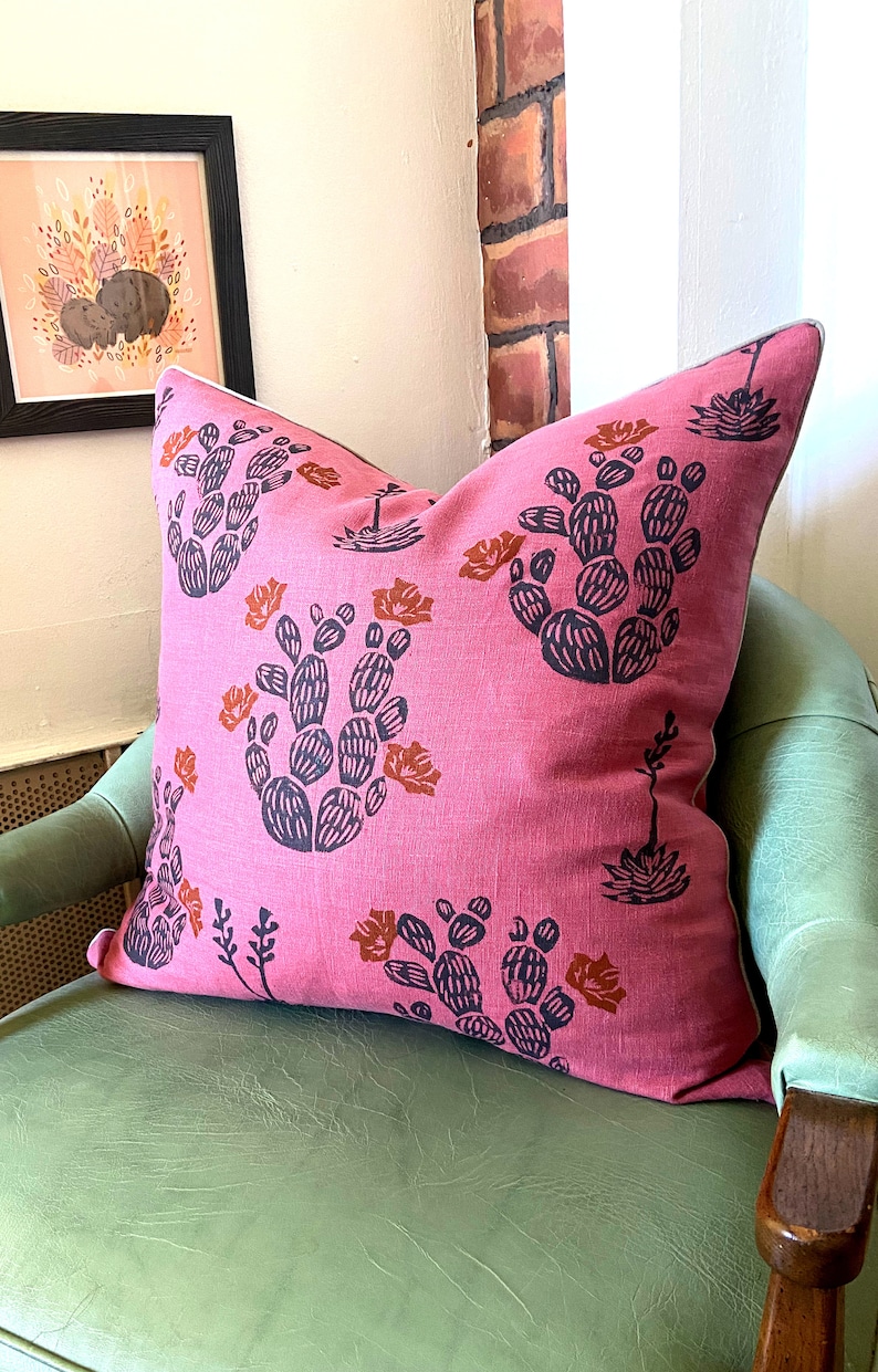 May include: A pink decorative pillow with a cactus pattern in navy blue and orange. The pillow is on a green chair, with a framed picture in the background.