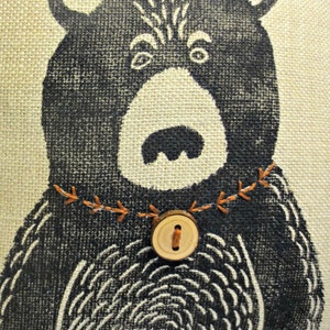 May include: A black and white illustration of a bear with a wooden button and orange stitching around the neck. The bear is on a beige burlap background.