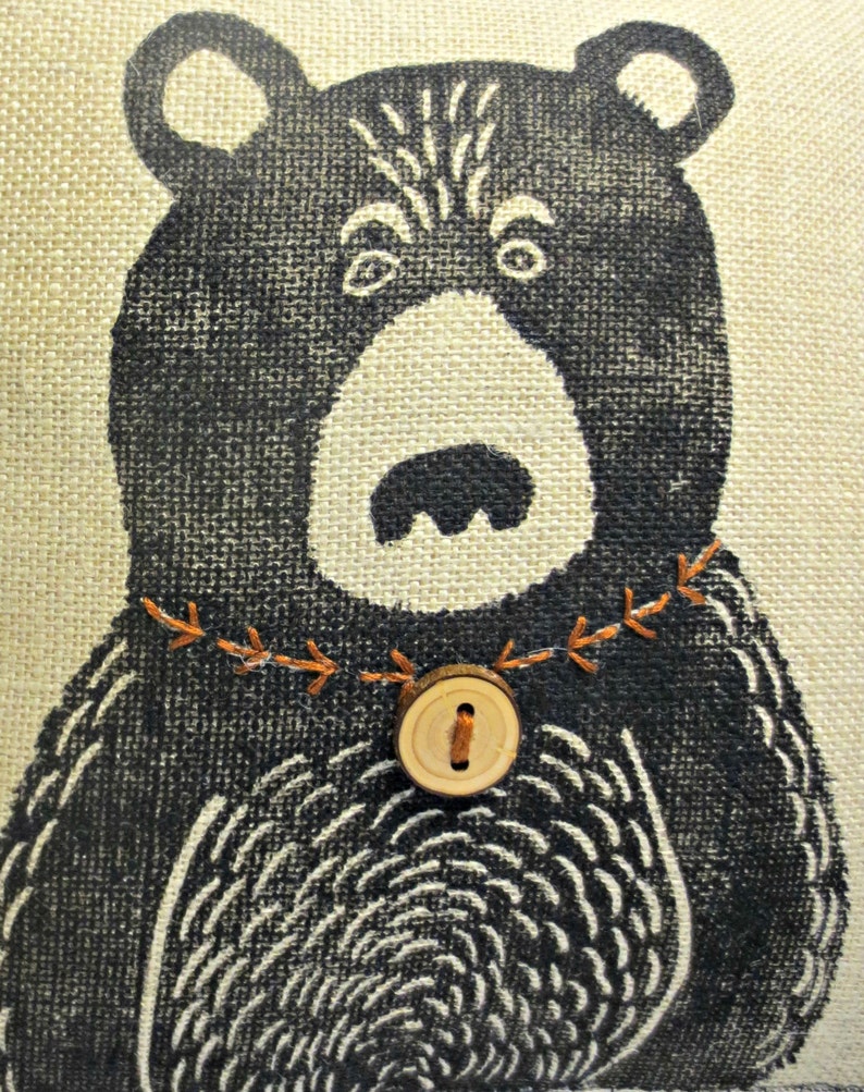 May include: A black and white illustration of a bear with a wooden button and orange stitching around the neck. The bear is on a beige burlap background.