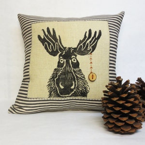 May include: Square cushion with a black and white moose design on a beige backdrop. It has a striped border and a wooden button detail. Two pine cones are in the foreground, adding a rustic touch.