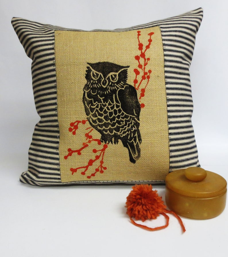 May include: A black and white owl with red berries printed on a burlap pillow cover. The pillow cover has a black and white striped border.