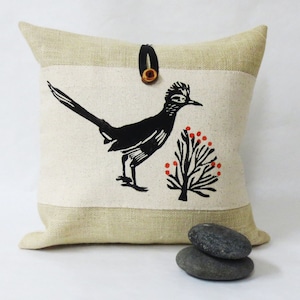 May include: A beige linen cushion with a black and white illustration of a roadrunner bird and a small tree with red berries. The cushion has a brown leather button closure.