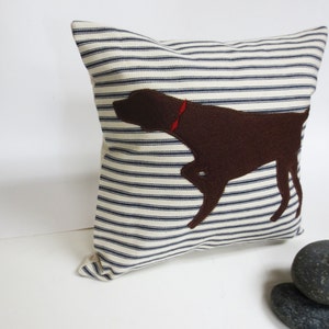May include: A white and blue striped cushion with a brown felt dog silhouette. The dog has a red collar.