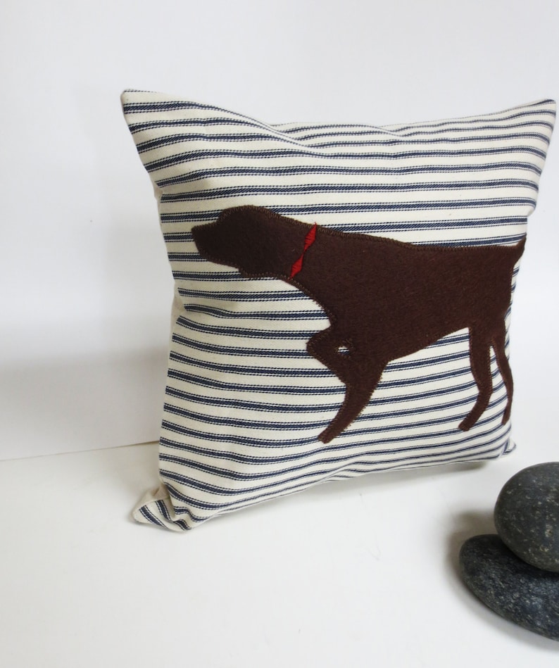 May include: A white and blue striped cushion with a brown felt dog silhouette. The dog has a red collar.