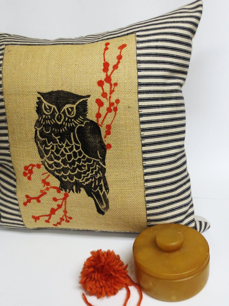 May include: A decorative pillow with a black and white owl design on a burlap background. The owl is perched on a branch with red berries. The pillow has a black and white striped border.