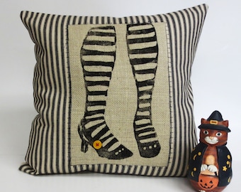 Op de afbeelding: Vierkantig kussen met een zwart-wit streepjespatroon. Het midden heeft een jute paneel met een zwart-wit gestreept been- en schoenontwerp. Een kleine gele knoop accentueert de schoen. Een keramieken kattenfiguur in een heksenkostuum staat naast het kussen.