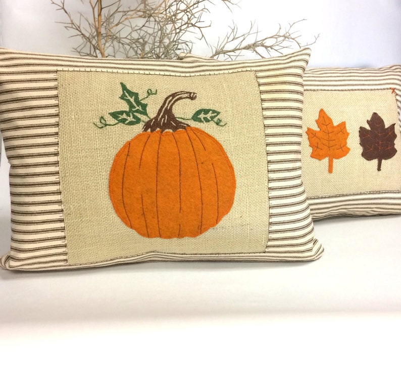 Op de afbeelding: Twee decoratieve kussens met een herfstthema. Een kussen is gemaakt van een bruin en wit gestreept stof met een jute patch in het midden. De jute patch heeft een oranje pompoen met een groene stengel en bladeren geborduurd. Het andere kussen is gemaakt van een bruin en wit gestreept stof met twee bruine en oranje bladeren geborduurd.