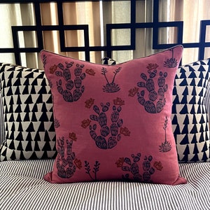 May include: A decorative pillow with a dusty rose background features a repeating pattern of dark blue cacti and rust-colored flowers. The pillow is bordered with a dark trim and is placed on a striped surface.