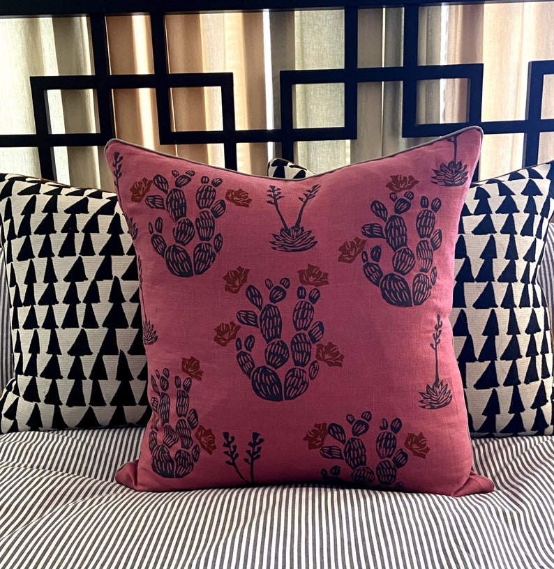 May include: A decorative pillow with a dusty rose background features a repeating pattern of dark blue cacti and rust-colored flowers. The pillow is bordered with a dark trim and is placed on a striped surface.