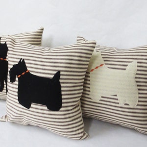 May include: Three decorative throw pillows with brown and white striped fabric. Two pillows feature black felt silhouettes of Scottish Terriers, and one pillow features a white felt silhouette of a West Highland White Terrier.