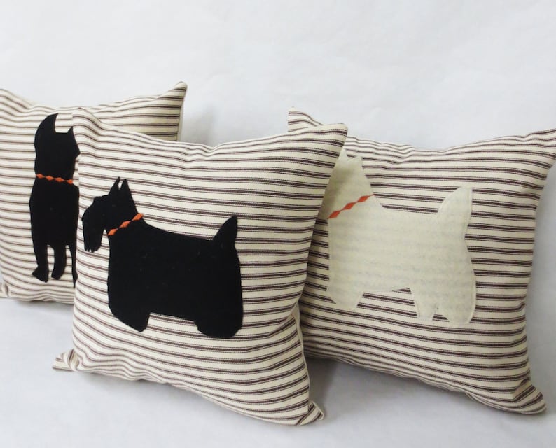 May include: Three decorative throw pillows with brown and white striped fabric. Two pillows feature black felt silhouettes of Scottish Terriers, and one pillow features a white felt silhouette of a West Highland White Terrier.