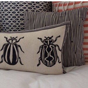 Puede incluir: Dos almohadas blancas con dise&ntilde;os de escarabajos negros. Las almohadas est&aacute;n sobre una cama blanca con una almohada estampada en blanco y negro detr&aacute;s de ellas.