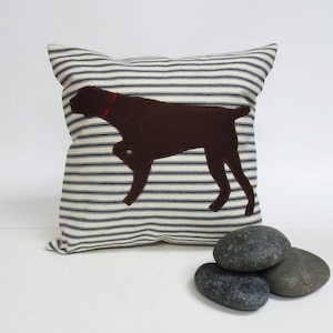 Könnte beinhalten: Ein braunes und weiß gestreiftes Kissen mit einer braunen Filzhund-Silhouette. Der Hund steht auf allen vieren und trägt ein rotes Halsband.