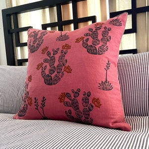 May include: A square, pink decorative pillow with a cactus print in black and brown. The pillow is on a striped bedspread. The pillow has a white trim.