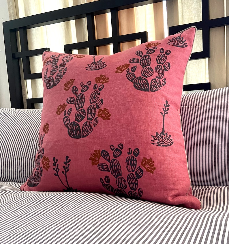 May include: A square, pink decorative pillow with a cactus print in black and brown. The pillow is on a striped bedspread. The pillow has a white trim.