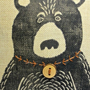 May include: A black and white illustration of a bear with a brown button and orange stitching around the neck on a beige burlap background.