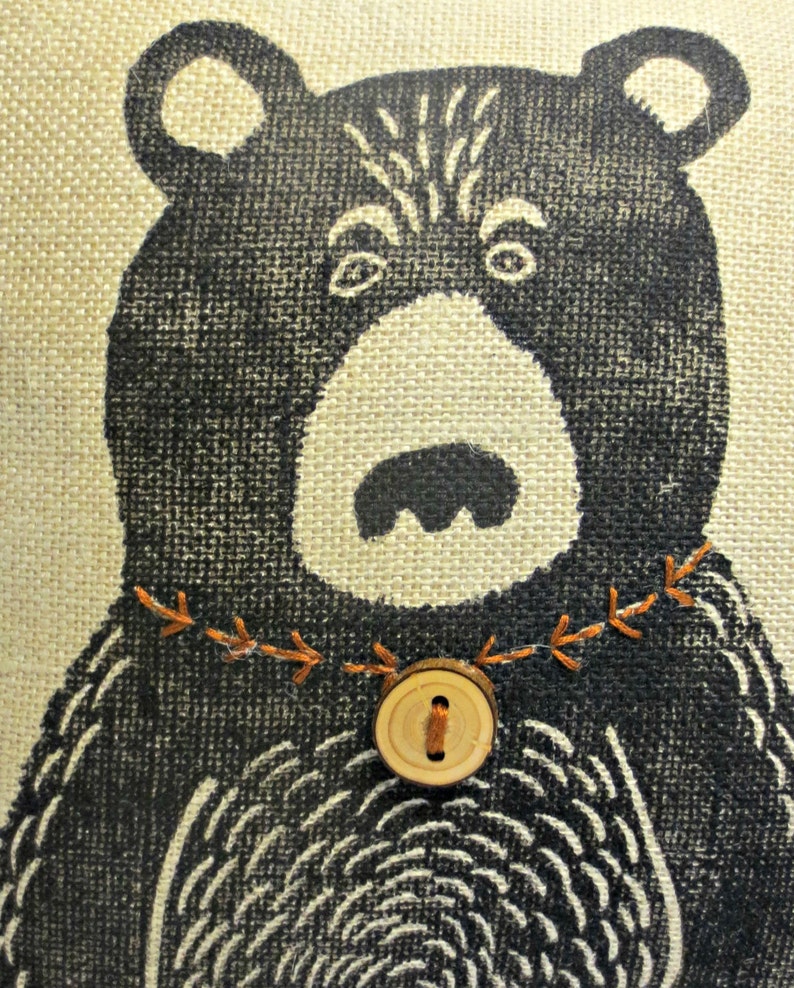 May include: A black and white illustration of a bear with a brown button and orange stitching around the neck on a beige burlap background.
