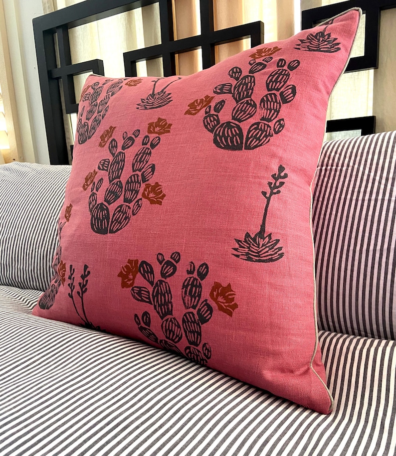 May include: A square decorative pillow with a pink background and a repeating pattern of black and brown cacti. The pillow has a white border and is resting on a striped pillow and a black headboard.