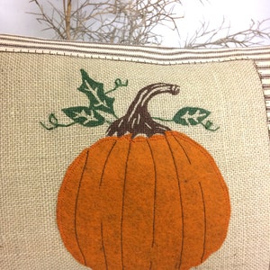 Op de afbeelding: Een bruine en beige jute kussen met een grote oranje viltpompoen met groene viltbladeren. De pompoen heeft een bruine viltstengel.