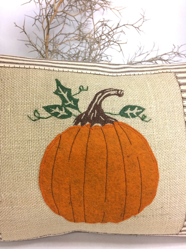 Op de afbeelding: Een bruine en beige jute kussen met een grote oranje viltpompoen met groene viltbladeren. De pompoen heeft een bruine viltstengel.