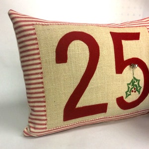 May include: A red and white striped pillow with a burlap front featuring the number 25 in red felt. A small green holly sprig with red berries is attached to the number 5.