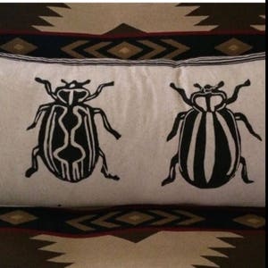 Puede incluir: Una almohada blanca con dise&ntilde;os de escarabajos en blanco y negro. Los escarabajos tienen un patr&oacute;n a rayas y est&aacute;n orientados en direcciones opuestas.