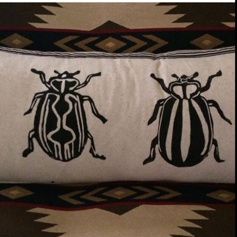 Puede incluir: Una almohada blanca con dise&ntilde;os de escarabajos en blanco y negro. Los escarabajos tienen un patr&oacute;n a rayas y est&aacute;n orientados en direcciones opuestas.