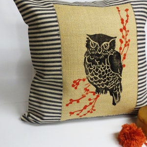 May include: A decorative pillow with a black and white owl printed on a burlap fabric background. The owl is perched on a branch with red berries. The pillow is surrounded by a black and white striped fabric.