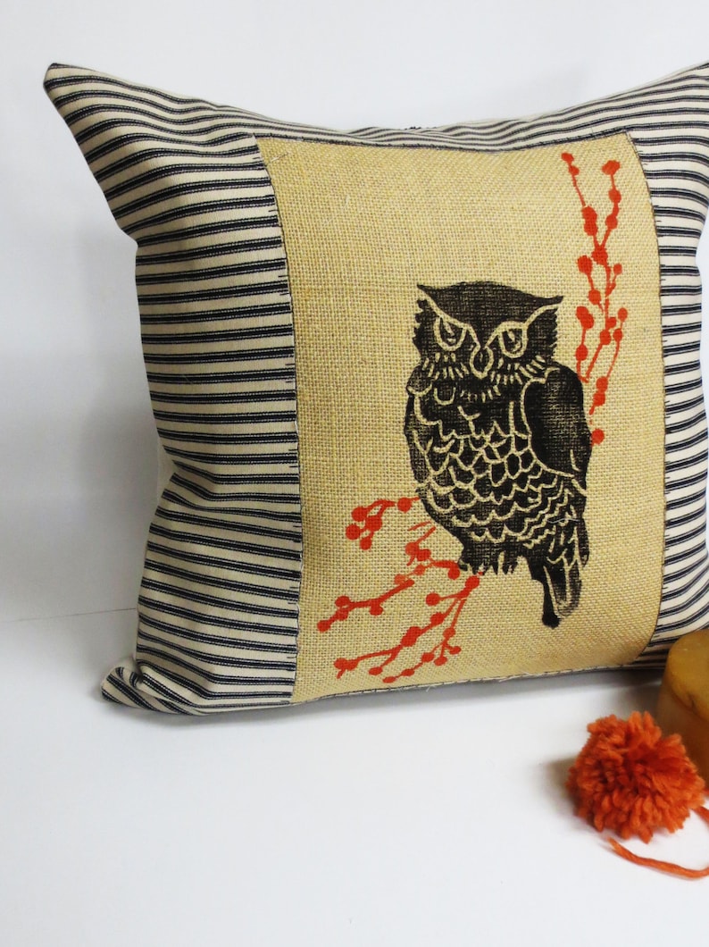 May include: A decorative pillow with a black and white owl printed on a burlap fabric background. The owl is perched on a branch with red berries. The pillow is surrounded by a black and white striped fabric.
