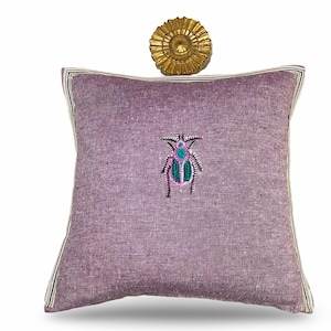 May include: A square, lavender-colored decorative pillow with an embroidered insect design in shades of pink, teal, and green. The pillow has a silver and white striped border and is topped with a gold-colored sunburst wall decor.