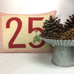 May include: A red and white striped pillow with the number 25 embroidered on it in red. The number is on a beige burlap background. A small green holly sprig is embroidered next to the number. The pillow is sitting on a white surface. Two pine cones are sitting on a silver metal cake stand.
