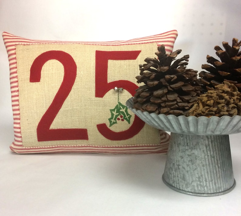 May include: A red and white striped pillow with the number 25 embroidered on it in red. The number is on a beige burlap background. A small green holly sprig is embroidered next to the number. The pillow is sitting on a white surface. Two pine cones are sitting on a silver metal cake stand.
