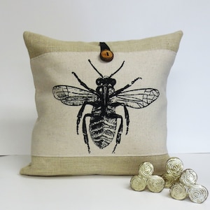 May include: A beige linen pillow with a black and white bee illustration and a brown button closure. The pillow has a burlap border.