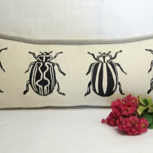 Puede incluir: Una almohada larga y rectangular con fondo blanco y dise&ntilde;os de escarabajos negros. La almohada tiene un borde rayado blanco y negro.