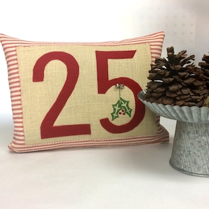 May include: Red and white striped pillow with the number 25 in red on a beige background. A small green holly sprig is attached to the number 5.