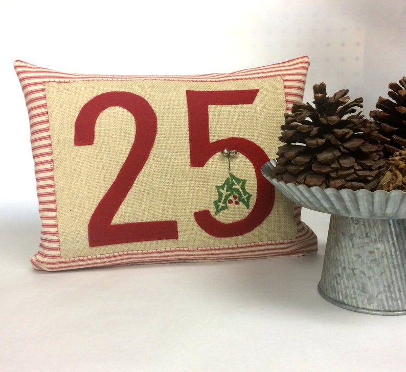 May include: Red and white striped pillow with the number 25 in red on a beige background. A small green holly sprig is attached to the number 5.