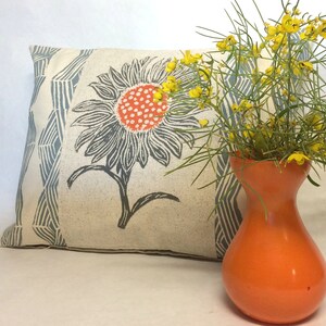 May include: A white pillow with a blue and gray geometric pattern and a black and white sunflower print. The pillow is sitting next to an orange vase with yellow flowers.