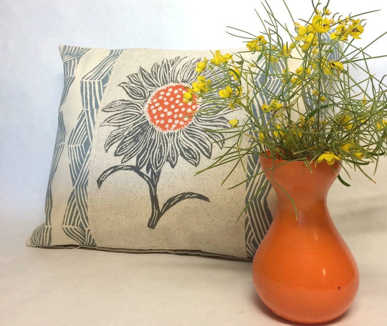 May include: A white pillow with a blue and gray geometric pattern and a black and white sunflower print. The pillow is sitting next to an orange vase with yellow flowers.