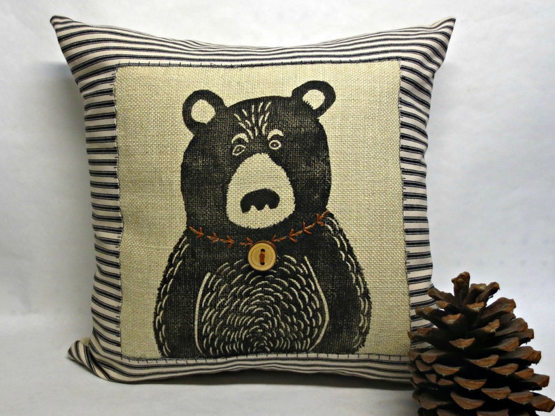 May include: A black and white pillow with a black bear design. The bear is wearing a brown twig necklace with a wooden button. The pillow has a white and black striped border.