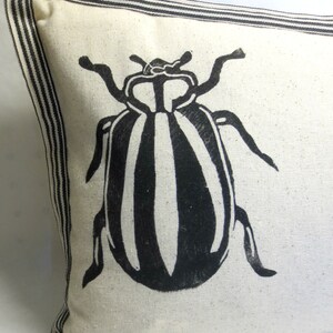 Puede incluir: Una almohada blanca con rayas negras y blancas alrededor de los bordes. La almohada tiene un gr&aacute;fico en blanco y negro de un escarabajo en la parte delantera.