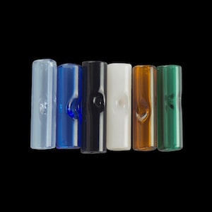 Puede incluir: Colección de siete tubos de vidrio de varios colores, incluyendo azul claro, azul oscuro, negro, blanco, ámbar y verde. Cada tubo tiene una hendidura redondeada. Los tubos están dispuestos sobre un fondo negro.