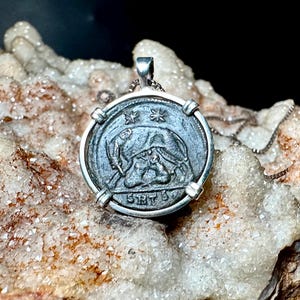 May include: A silver pendant necklace featuring a circular coin design. The coin has an embossed image of a figure and text. The pendant is attached to a delicate chain. The background is a light-colored, textured surface.