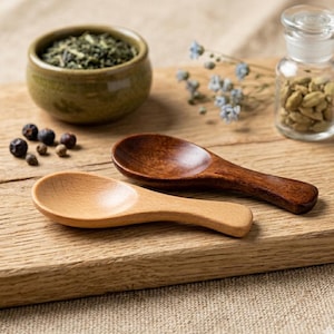 Peut inclure: Deux cuillères en bois, une claire et une foncée, sont posées sur une planche en bois. Un petit bol en céramique rempli d'herbes vertes, un bocal en verre avec des graines et des grains de poivre sont également présents. Les cuillères sont pour les épices.