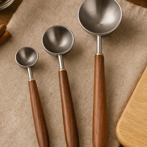 Puede incluir: Tres cucharas medidoras de acero inoxidable con mangos de madera. Las cucharas están dispuestas de menor a mayor, con un cuenco plateado y un mango liso de color marrón oscuro. Una banda dorada conecta el cuenco con el mango.