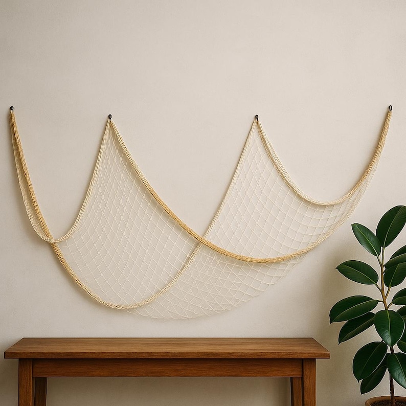 Puede incluir: Una red de pesca decorativa de color blanquecino cuelga de una pared neutra. Est&aacute; asegurada por tres ganchos negros. Debajo hay un banco de madera y a la derecha una planta en maceta con hojas verdes.