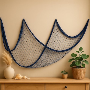 Może przedstawiać: Granatowa dekoracyjna sieć rybacka wisi na beżowej ścianie. Sieć jest ułożona w falisty wzór. Drewniany stół poniżej prezentuje wazon z suszonymi roślinami, muszlami i roślinami doniczkowymi, tworząc motyw marynistyczny.
