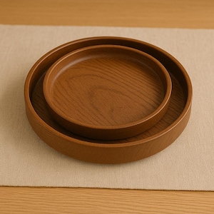 Op de afbeelding: Twee houten serveerschalen die in elkaar passen. De schalen hebben een warme bruine kleur met zichtbare houtnerf. De grotere schaal heeft een diameter van ongeveer 25 cm, terwijl de kleinere schaal ongeveer 15 cm in diameter is. De schalen liggen op een beige ondergrond.