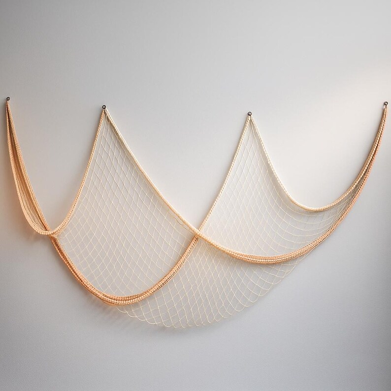 Puede incluir: Una red de pesca decorativa, de color crema con un borde marr&oacute;n claro, cuelga contra una pared gris claro. La red est&aacute; suspendida de tres puntos, creando un patr&oacute;n drapeado en forma de ola. Esta decoraci&oacute;n de tem&aacute;tica n&aacute;utica a&ntilde;ade un toque costero.