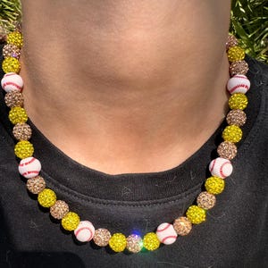 May include: A necklace made of yellow and brown beads with white baseballs with red stitching. The necklace is worn by a person with a black shirt.